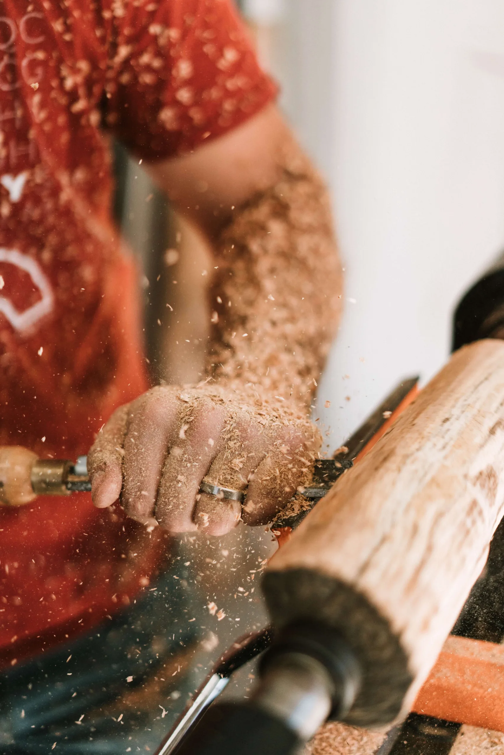 Man in een rood-wit T-shirt die een groot houten deegrol-achtig object/handgereedschap vasthoudt, gefotografeerd tegen een neutrale achtergrond