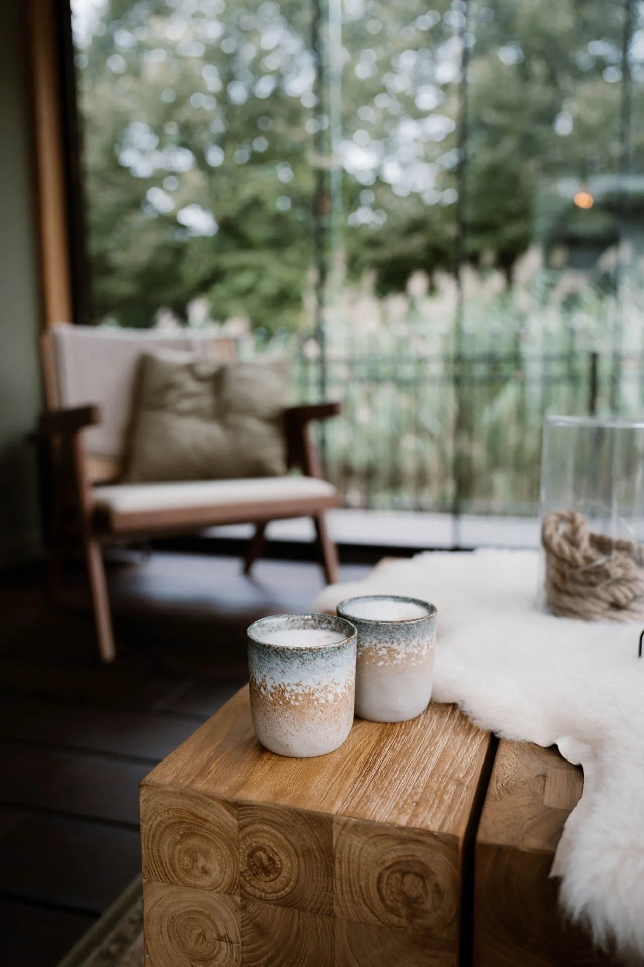 Twee keramische mokken op een houten bijzettafel in een sfeervolle lounge- of zithoek van een modern buitenverblijf met kussens en groen in de achtergrond.