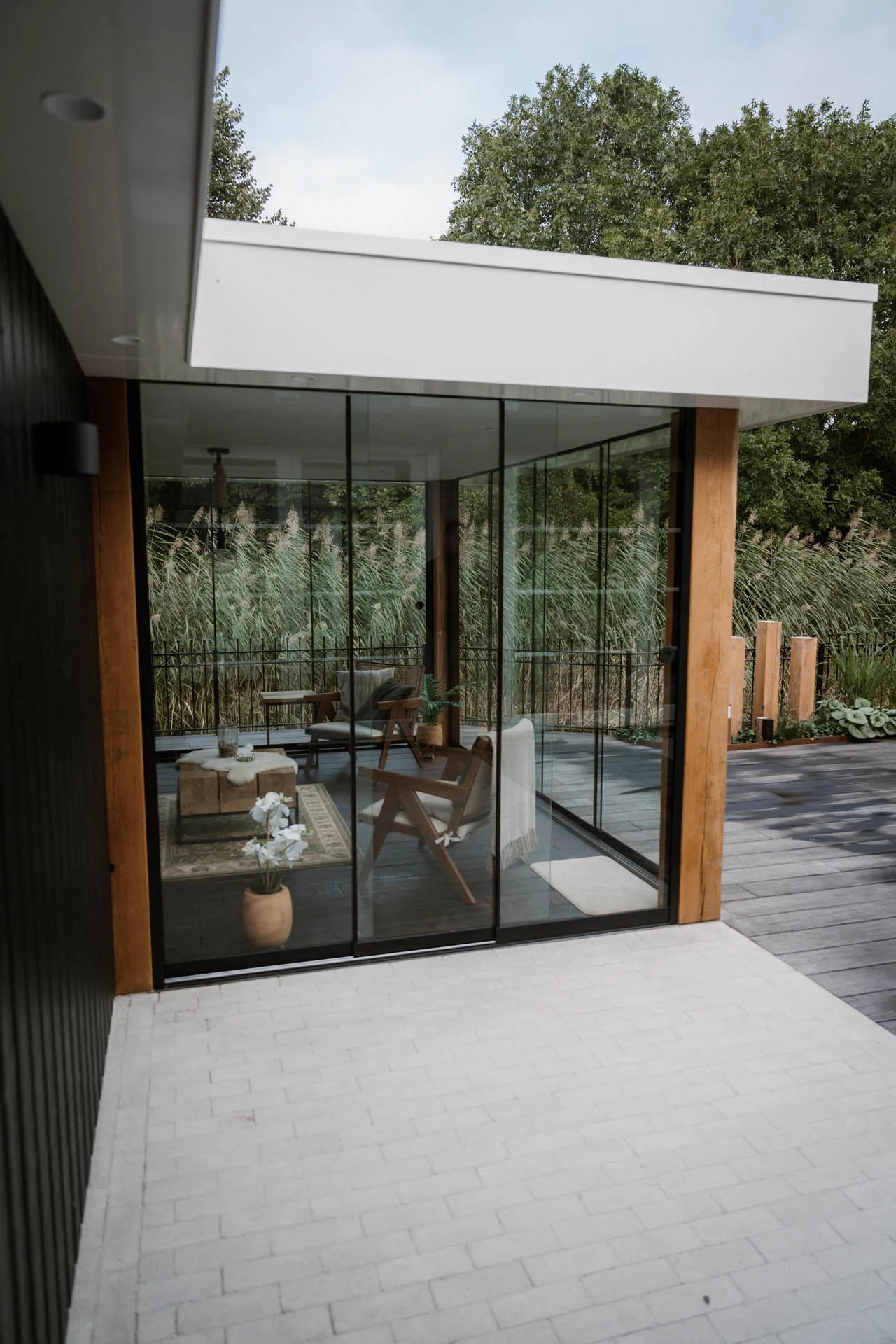Modern buitenverblijf met glazen schuifdeuren en houten kolommen, zicht op een zithoek binnen en een terrasomgeving met groene beplanting.