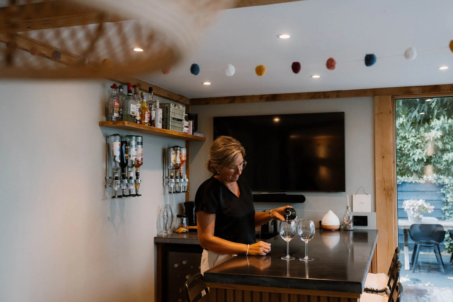 Vrouw die een glas wijn inschenkt aan een bar in een stijlvolle binnenruimte met planken vol drankflessen aan de muur, televisiescherm en grote glazen schuifdeur naar buiten.