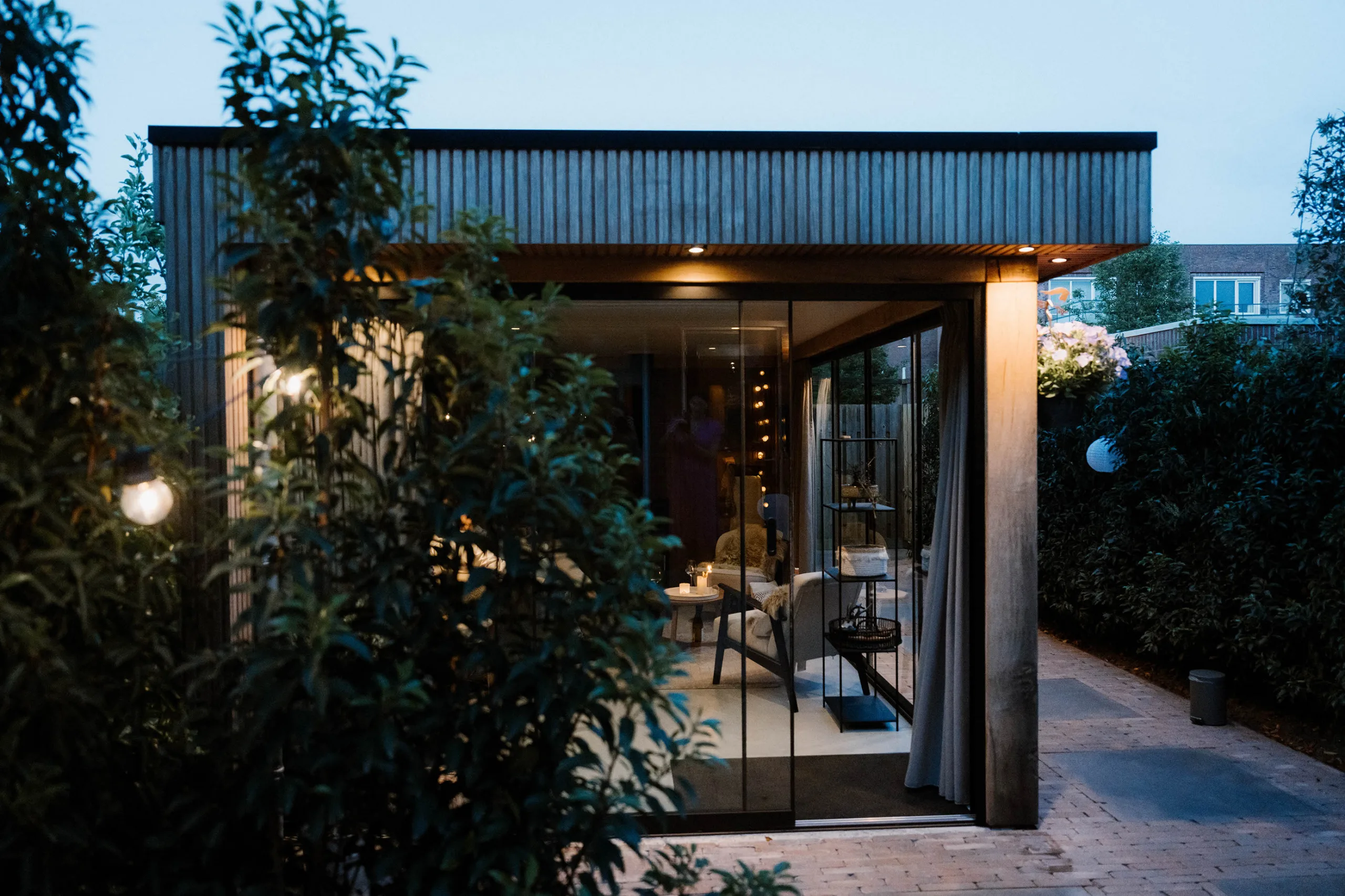 Avondbeeld van een modern buitenverblijf met houten gevelbekleding en grote glazen schuifdeuren, binnen verlichte zithoek zichtbaar, omgeven door groene beplanting.