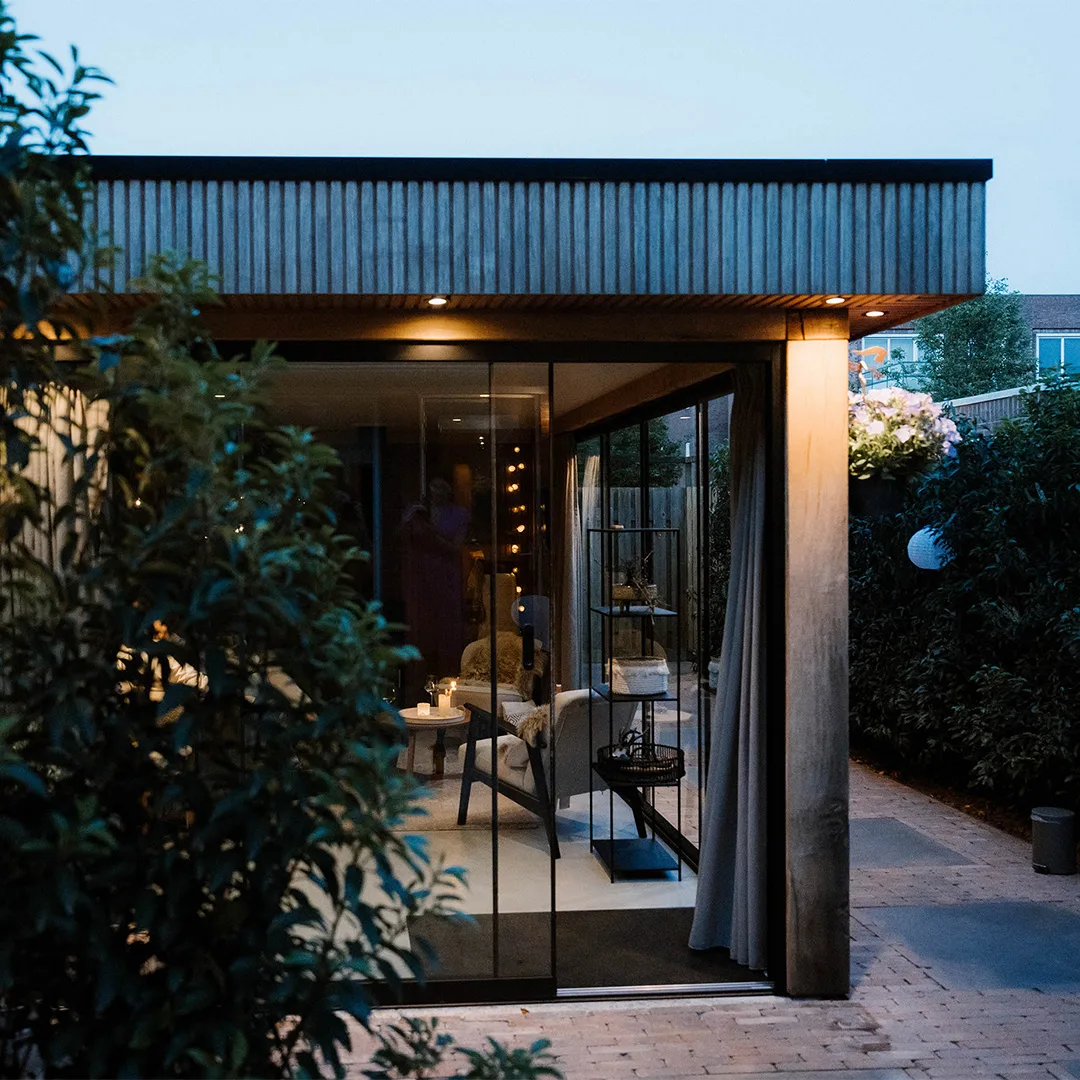 Avondbeeld van een modern buitenverblijf/tuinkamer met houten gevelbekleding en glazen schuifdeuren, waar binnen sfeervol verlichte zithoek zichtbaar is te midden van tuinplanten