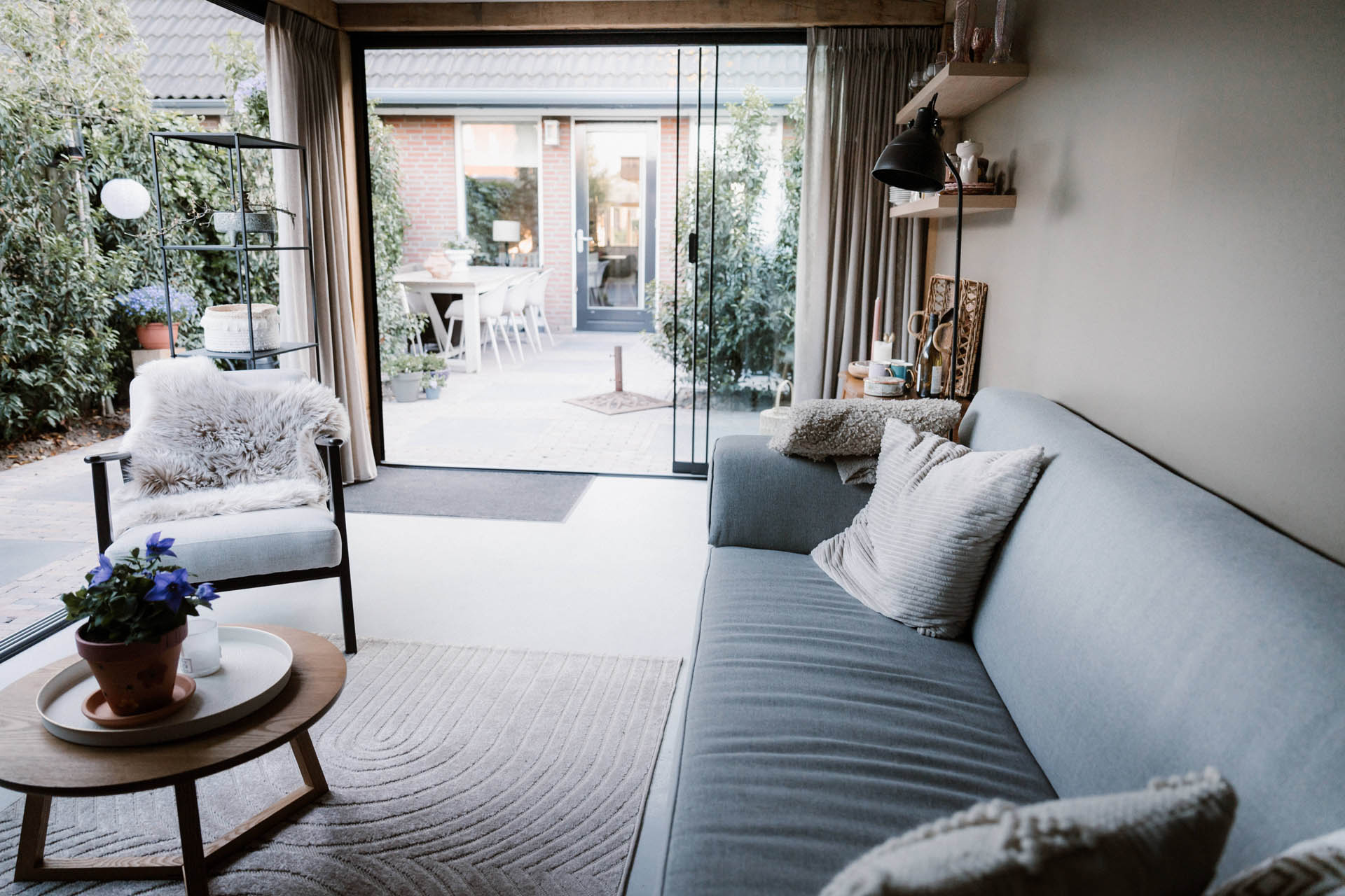 Gezellige en lichte zithoek in een woonruimte met een comfortabele bank, een fauteuil met zachte kussens, een lage salontafel met planten, en grote glazen schuifdeuren die uitkomen op een terras met tuinmeubilair.